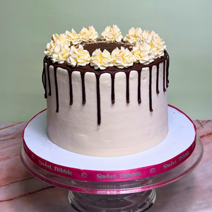 Decorative tres leches cake with white frosting, chocolate drizzle, and yellow flowers on a Sweet Nibble stand.